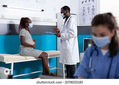 Pregnant woman talking to doctor at checkup appointment in medical office while wearing face masks. Physician doing healthcare consultation for patient with pregnancy expecting child.