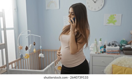 Pregnant woman talking on phone in baby room standing next to cradle surrounded by soft pastel colors and baby toys while natural light fills the cozy interior.