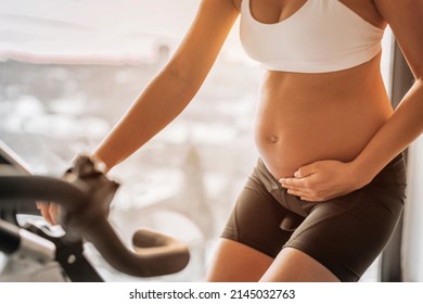 Pregnant Woman Training Cardio On Exercise Spin Bike At Indoor Fitness Gym Or Home Training On Stationary Bicycle. Closeup Of Fit Girl Holding Pregnancy Belly