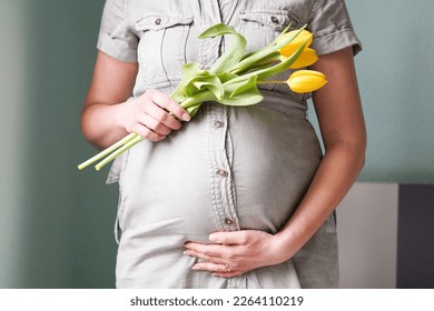 Pregnant woman with tulips flowers holding hands on belly on a pastel background. Mothers day or motherhood concept.
