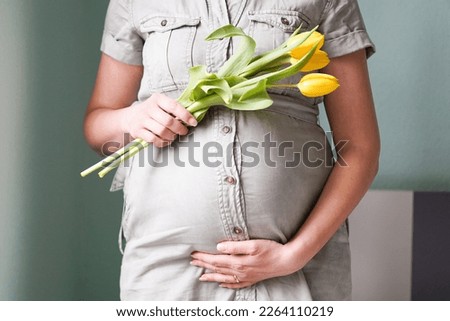 Pregnant woman with tulips flowers holding hands on belly on a pastel background. Mothers day or motherhood concept.