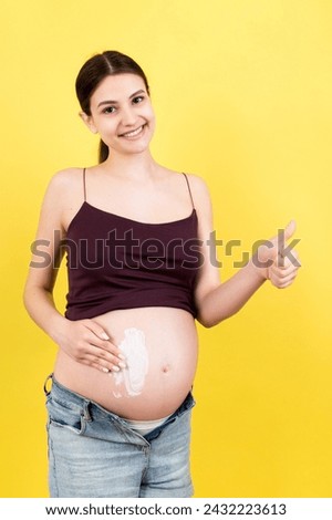 pregnant woman in unzipped jeans applying moisturizing cream on her belly against stretch marks at colorful background with copy space. Skin care concept.
