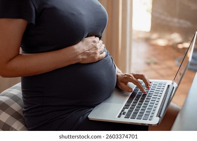 Pregnant woman using laptop, hands on belly, working from home. Technology and pregnancy, female multitasking, remote work. Relaxed home setting, casual attire. Pregnancy and maternity image.