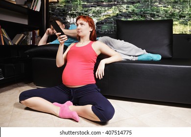 Pregnant Woman Watching TV Sitting On The Floor While Guy Laying Down On Couch Behind