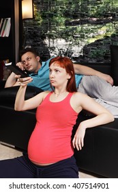 Pregnant Woman Watching TV Sitting On The Floor While Guy Laying Down On Couch Behind Closeup