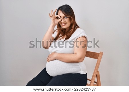 Pregnant woman wearing band aid for vaccine injection smiling happy doing ok sign with hand on eye looking through fingers 