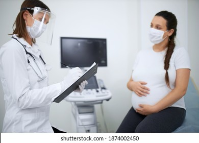 Pregnant woman wearing medical face mask during visit at modern clinic. Female doctor in protective mask doing regular check up for patient. Concept of pandemic and motherhood.
