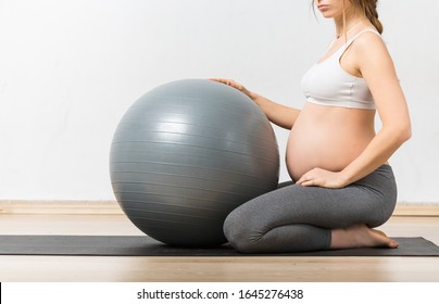 Pregnant woman wearing yoga pants, gym clothing, sitting with legs bent on a yoga mat on wooden hard floor, holding a Pilates ball with right hand and her hip with her left hand