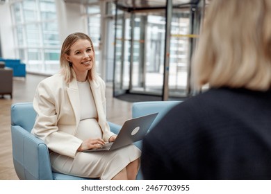 Pregnant woman in white suit at business meeting with colleague in a modern office space or coworking center. Professional businesswoman multitasking with laptop during work discussion. - Powered by Shutterstock