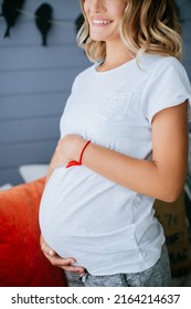 Pregnant Woman In A White T-shirt And Gray Shorts Holding Her Belly, Kneeling On The Couch And Smiling (Authoring Photo)