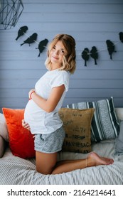 Pregnant Woman In A White T-shirt And Gray Shorts Holding Her Belly, Kneeling On The Couch And Smiling (Authoring Photo)