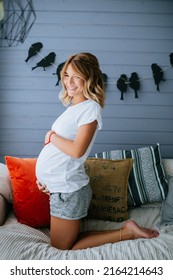 Pregnant Woman In A White T-shirt And Gray Shorts Holding Her Belly, Kneeling On The Couch And Smiling (Authoring Photo)