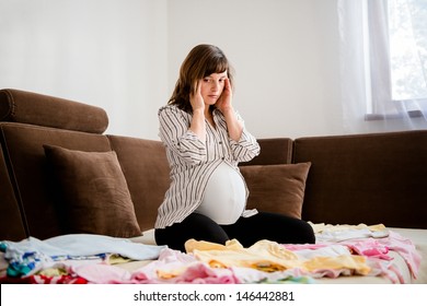 Pregnant Woman Worries Above Baby Clothes At Home On Sofa