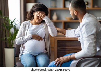 Pregnant Young Black Woman Feeling Bad, Sitting On Chair At Clinic, Touching Her Big Tummy And Head, Visiting Doctor, African American Man Gynecologist Comforting Crying Expecting Lady