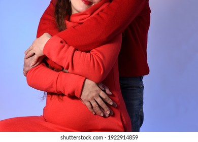 A Pregnant Young Woman In A Red Knitted Dress, Being Hugged By Her Husband Standing Behind Her. A Hand Rests On Her Belly. A Smile Can Be Seen On Her Lips. Expecting A Baby. Blue Background. Studio. 