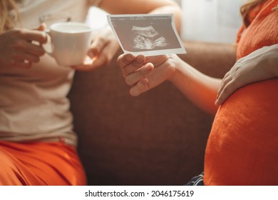 pregnant young woman sitting in kitchen with girlfriend showing photo card of ultrasound snapshot to sister, life pregnancy concept