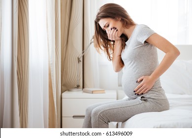 Pregnant young woman sitting on bed and feeling sick at home