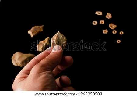 a prehistoric man holds a stone age borer in his hand. paleolithic period. black background. in the background you see the ostrich eggshell beads to make a necklace