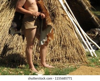 Prehistoric Man In Loincloth And Animal Skins Kindles The Fire Near The Straw Hut. Historical Reconstruction Of Neanderthal And Cro-magnon Life