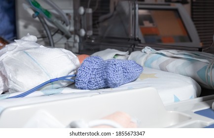 Premature Newborn Recovering In Incubator. His Foot In A Blue Sock In Focus, With A Premature Infant Pulse Oximeter. Intensive Care Unit, Ventilator For Artificial Lung Ventilation As The Background