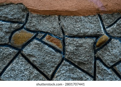 Premium full-frame texture shot showcasing a rustic stone masonry wall topped with a vibrant architectural border. The horizontal composition features split-face grey granite blocks with a coarse salt-and-pepper texture, beautifully outlined by thick, dark charcoal or black painted mortar joints that accentuate the geometric mosaic layout. Artfully nestled within the grey stones are a few irregular accent rocks in warm ochre, gold, and earthy brown tones, creating an eye-catching color contrast. At the top, a solid stripe of textured, terracotta-orange painted stucco completes the dynamic layer. This powerful graphic combination of raw stone masonry and bold color blocks is an exceptional design asset engineered for high-end digital texturing, 3D mapping, contemporary web design backgrounds, and rustic real estate branding.