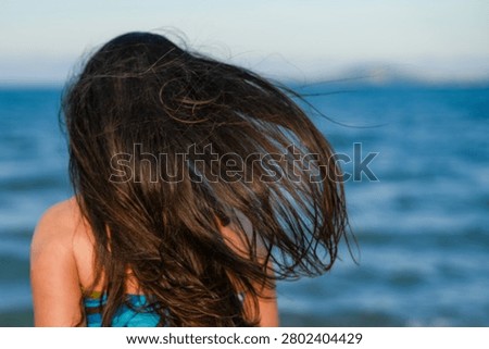 Premium medium close-up shot from behind showcasing a brunette woman's long, dark brown hair blowing dramatically in the sea breeze. The horizontal composition features a dynamic selective focus on the beautiful, windswept texture of the hair strands, captured mid-movement during a sunset or sunrise. The background displays a soft, artistic bokeh of the vibrant blue ocean waters stretching toward a distant coastline under a clear sky. The image evokes strong feelings of freedom, wellness, summer vacation adventures, and natural hair care. This high-quality lifestyle visual asset is exceptionally engineered for beauty and hair salon marketing, travel agency promotions, mental wellness blogs, and summer-themed editorial layouts.

