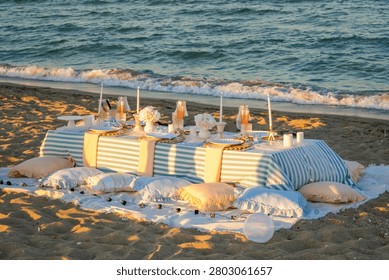 Premium wide-angle shot capturing an elegant, romantic picnic setup on a sandy beach during the warm golden hour, arranged in a balanced horizontal composition. The scene features a low table draped in a blue and white striped tablecloth, beautifully adorned with tall white candles, delicate wine glasses, and pastel pink tableware. Soft pillows, cushions, and fluffy blankets are scattered around the white base rug, flanked by glowing decorative light globes. The low-angle evening sun casts long, dramatic shadows across the textured sand dunes in the foreground, while large coastal tetrapods line the background. This high-quality lifestyle asset is exceptionally engineered for luxury event planning promotions, romantic beach travel editorials, summer wedding inspiration, and elegant outdoor dining marketing campaigns.
