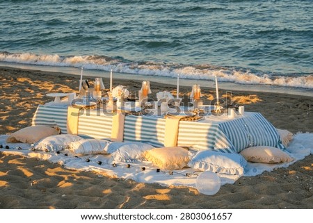 Premium wide-angle shot capturing an elegant, romantic picnic setup on a sandy beach during the warm golden hour, arranged in a balanced horizontal composition. The scene features a low table draped in a blue and white striped tablecloth, beautifully adorned with tall white candles, delicate wine glasses, and pastel pink tableware. Soft pillows, cushions, and fluffy blankets are scattered around the white base rug, flanked by glowing decorative light globes. The low-angle evening sun casts long, dramatic shadows across the textured sand dunes in the foreground, while large coastal tetrapods line the background. This high-quality lifestyle asset is exceptionally engineered for luxury event planning promotions, romantic beach travel editorials, summer wedding inspiration, and elegant outdoor dining marketing campaigns.
