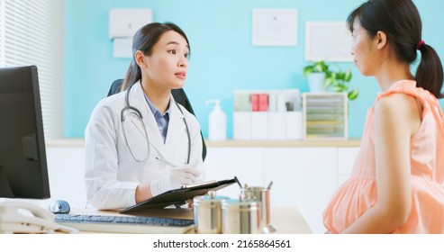 Prenatal Exam Concept - Asian Female Doctor Wearing White Coat Is Diagnosing Pregnant Woman And She Takes Note On Paper Document In Hospital