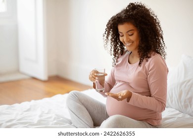 Prenatal Vitamins. Beautiful Smiling Black Pregnant Woman Holding Pills And Glass Of Water, African American Expectant Mother Taking Supplements For Healthy Pregnancy, Sitting On Bed At Home - Powered by Shutterstock