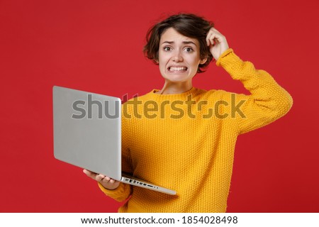 Preoccupied puzzled young brunette woman 20s in basic casual yellow sweater standing working on laptop pc computer put hand on head looking camera isolated on bright red background studio portrait