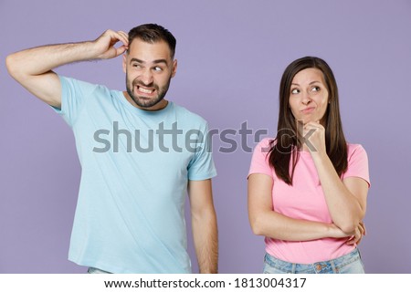 Preoccupied puzzled young couple two friends man woman 20s in blue pink empty blank t-shirts put hand prop up on chin head looking aside isolated on pastel violet color background studio portrait