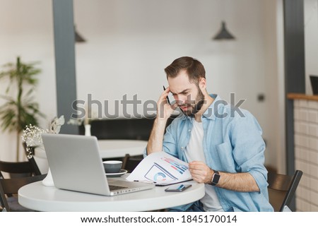 Preoccupied puzzled young man sitting alone at table in coffee shop cafe restaurant indoors working or studying on laptop pc computer with papers document. Freelance mobile office business concept