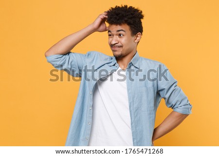 Preoccupied young african american guy in casual blue shirt posing isolated on yellow wall background studio portrait. People lifestyle concept. Mock up copy space. Put hand on head, looking aside