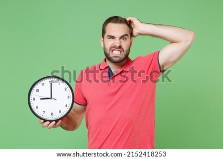 Preoccupied young bearded man guy in casual red pink t-shirt posing isolated on green background studio portrait. People emotions lifestyle concept. Mock up copy space. Hold clock, put hand on head