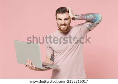 Preoccupied young bearded tattooed man guy in pastel casual t-shirt posing isolated on pink background studio. People lifestyle concept. Mock up copy space. Hold laptop pc computer put hand on head