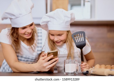 Preparation For Baking Of Mother And Child, Kid. Cute Daughter Hold Cooking Spatula In Hands And Look At Mobile Phone, Searching Recipe Of Cake