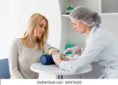Preparation for blood test with pretty young blond woman by female doctor in white coat medical uniform on the table in white bright room. Nurse pierces the patient's arm vein with needle blank tube.