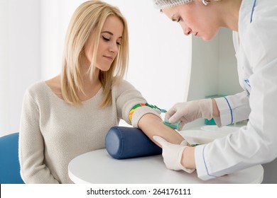 Preparation for blood test with pretty young blond woman by female doctor in white coat medical uniform on the table in white bright room. Nurse pierces the patient's arm vein with needle blank tube.