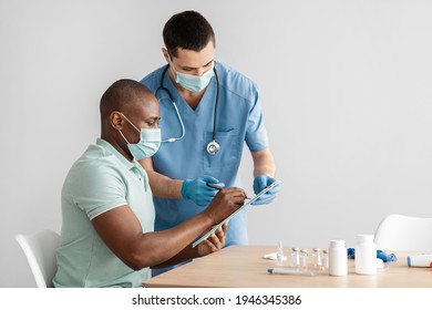 Preparation For Coronavirus Immunization And Health Care During Pandemic. Young Doctor In Protective Mask And Adult African American Patient Fill Out Questionnaire For Procedure At Table, Empty Space