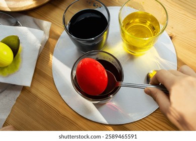 Preparation for Easter - the process of painting Easter eggs. Three glasses with different colors, a hand holding a painted egg on a spoon. Family traditions.