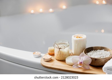Preparation for hotel spa treatment or home bath procedure. White washbasin in bathroom with accessories on tray. Burning candles, washcloth, soap, foot brush, bottle with sea salt, orchid flower