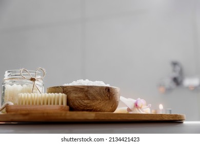 Preparation for hotel spa treatment or home bath procedure. White washbasin in bathroom with accessories on tray. Burning candles, soap, foot brush, glass bottle with sea salt, orchid flower