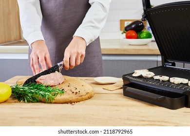 Preparation On Electric Grill. Woman In White Shirt And Apron Standing And Cutting Meat On The Kitchen.