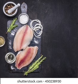 preparation of raw tilapia fillets with salt, oil and herbs, whole pepper in a mortar, laid out on a chopping board on a dark rustic background Border with space for text top view