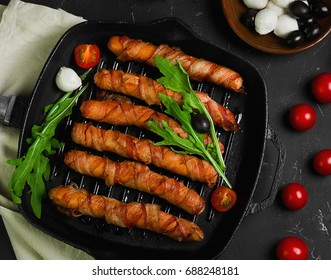 Preparation Of Roast Grill Sausages Wrapped Spirally In Bacon On A Cast-iron Frying Pan. Ingredients For Sausages In Bacon Cherry Tomatoes, Rucola Salad, Pepper. Black Concrete Background. Top View.