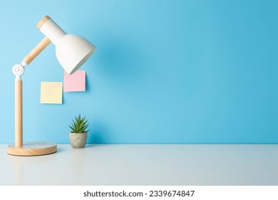 Prepare for classes with this side view picture of a tidy desk featuring desk lamp and tiny flowerpot. Empty blue wall with sticky notes ready for text or promotion