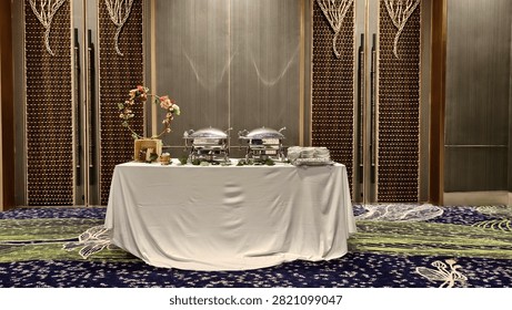 Prepared buffet table with covered chafing dishes, stacked plates, floral decoration, and serving equipment in an elegant banquet hall.
