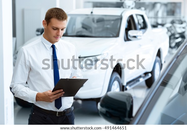 Preparing all\
info needed. Handsome male car dealer writing on his clipboard\
while working at the car salon\
copyspace