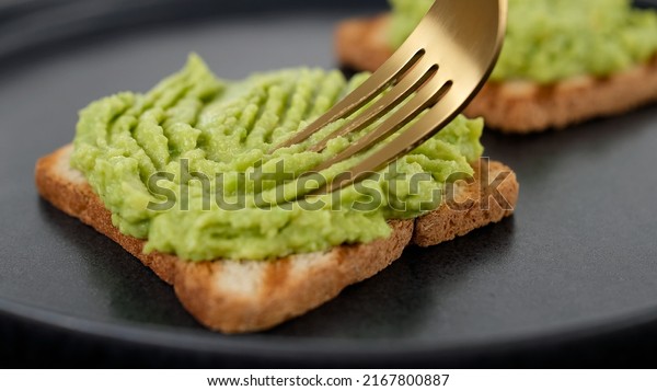 Preparing avocado toast.\
Hands with fork spreading mashed avocado on toasted whole  bread.\
Vegan food concept